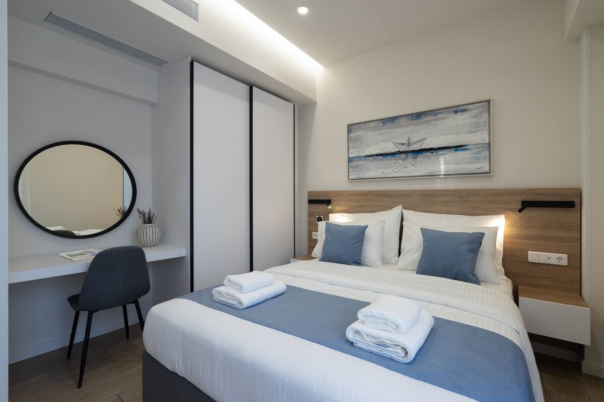 A modern hotel room with a large bed, bedside tables, a desk with a chair, a round mirror, and a painting on the wall.