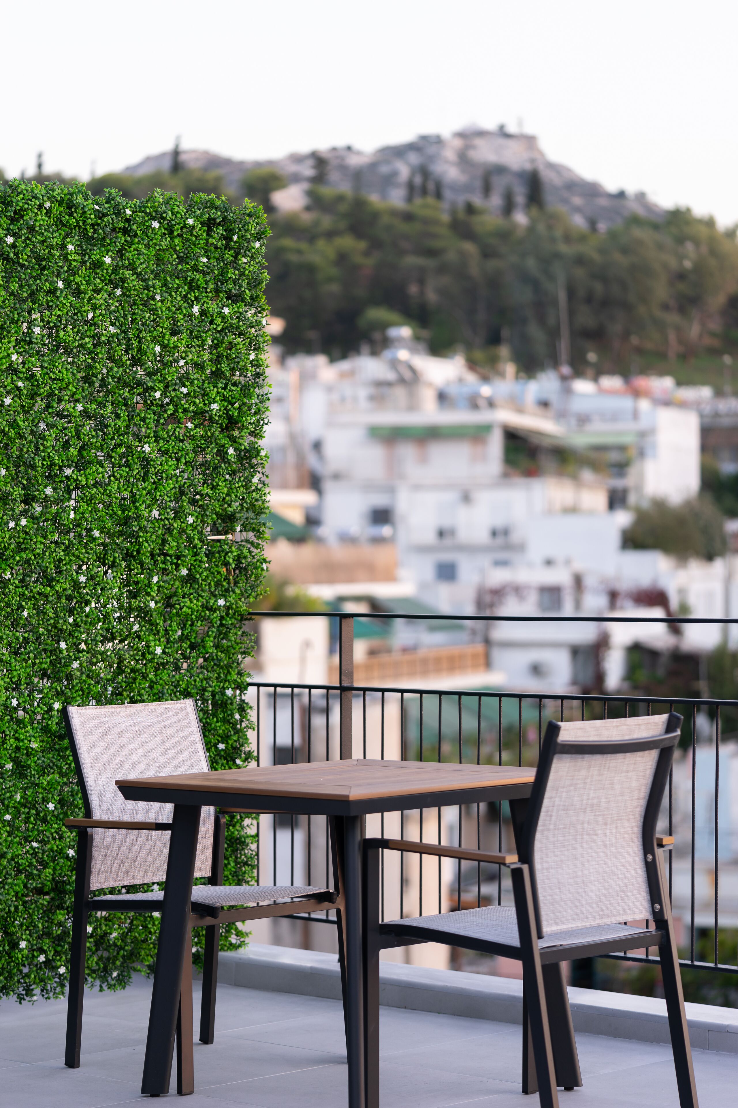 A balcony with a table and chairs, a green hedge, and a cityscape view in the background.