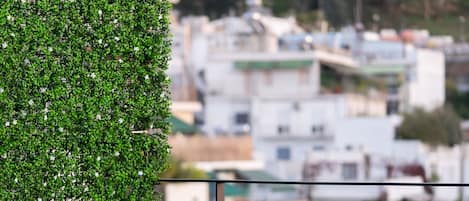 A balcony with a table and chairs, a green hedge, and a cityscape view in the background.