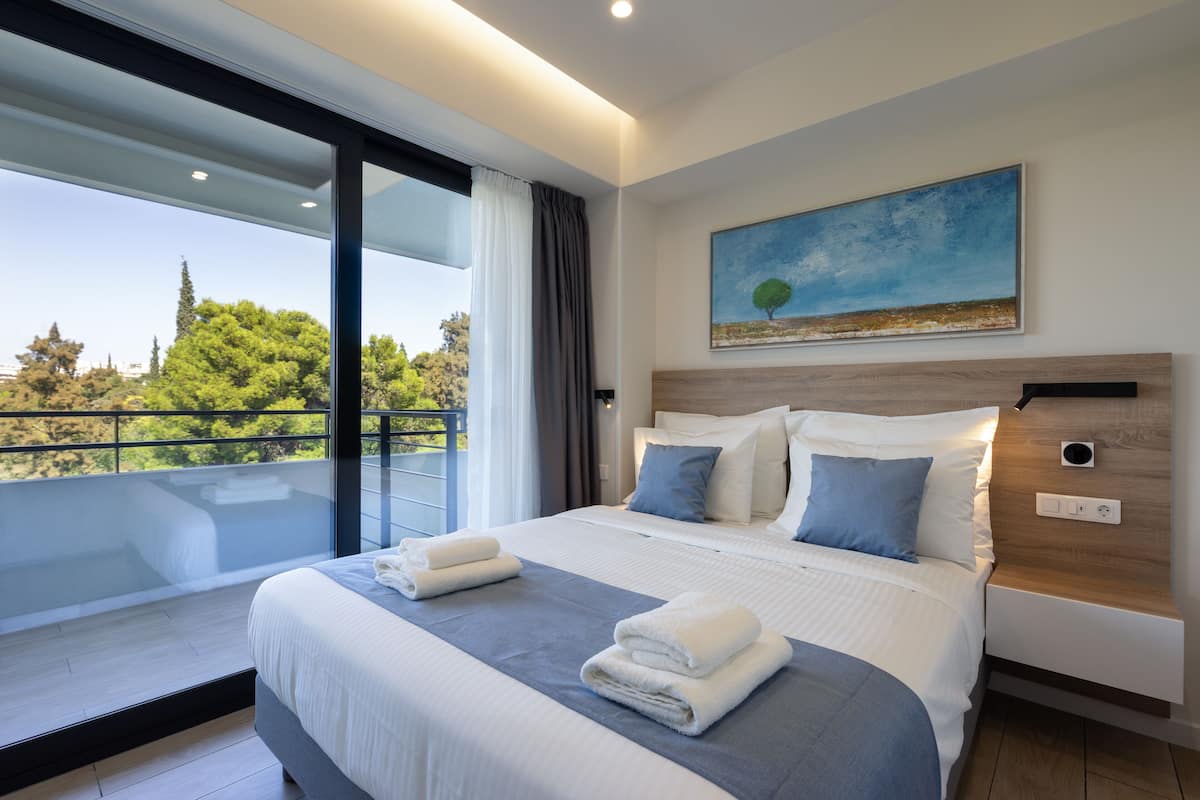 A modern hotel room with a large bed, a painting, and a balcony with a view of trees.