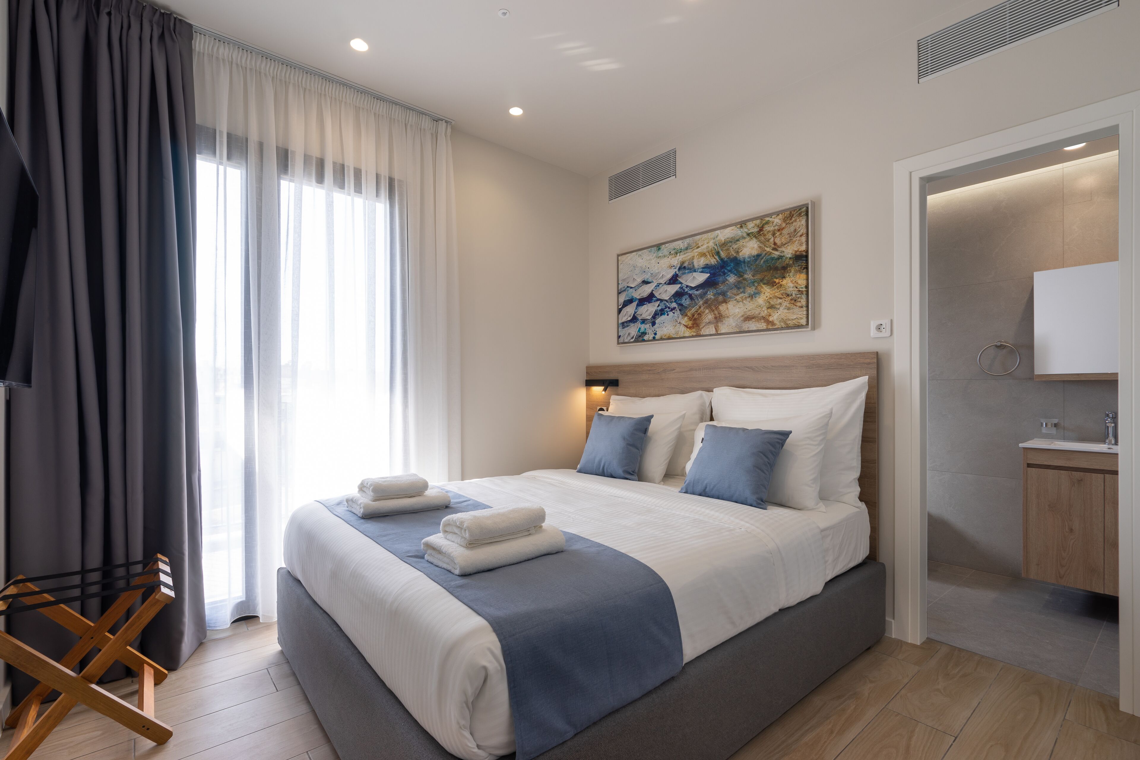 A modern hotel room with a large bed, a wooden headboard, a bedside table, a nightstand, a bathroom with a sink and mirror, and a window with curtains.