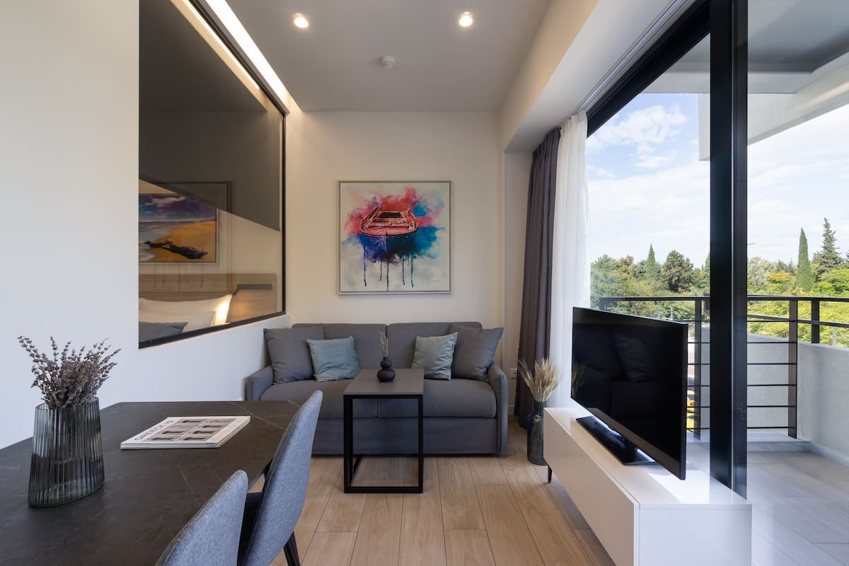 A modern living room with a grey sofa, a coffee table, and a flat-screen TV. There is a painting on the wall and a window with a view of trees.