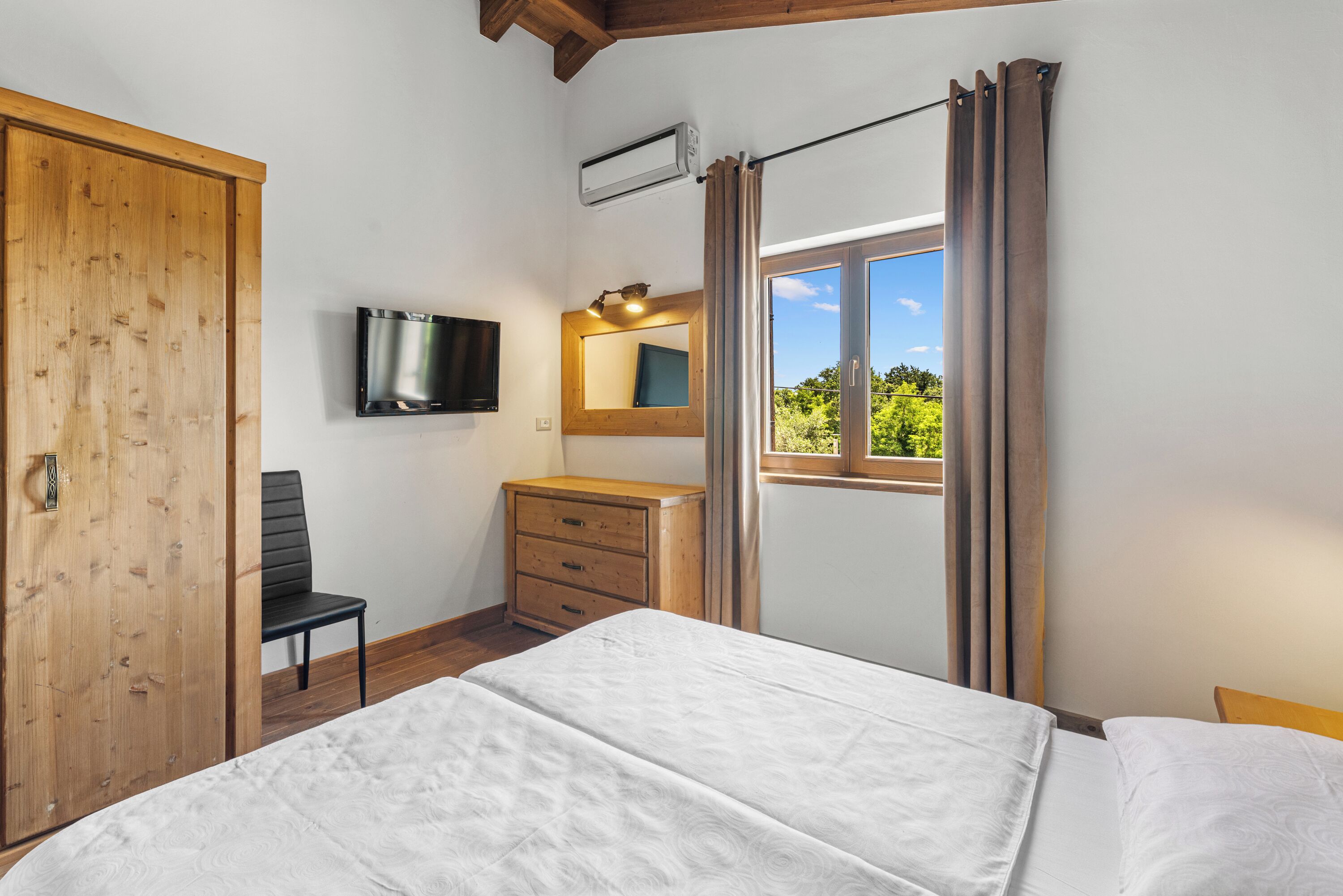 A bedroom with a bed, a wooden wardrobe, a TV mounted on the wall, a small desk with a chair, and a window with curtains.