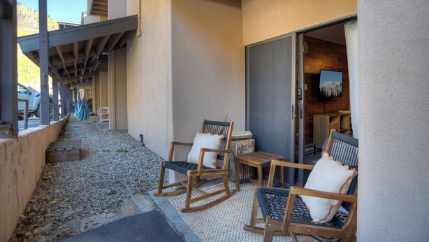 A covered outdoor seating area with wooden rocking chairs and a small table, a wooden bench, and a door leading to an interior space with a mounted TV. - Cozy Bear Den (#109) | Close to Lifts, Fireplace, Patio, No Stairs, Hot Tub (Durango)