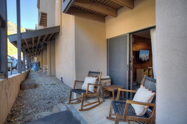 A covered outdoor seating area with wooden rocking chairs and a small table, a wooden bench, and a door leading to an interior space with a mounted TV.