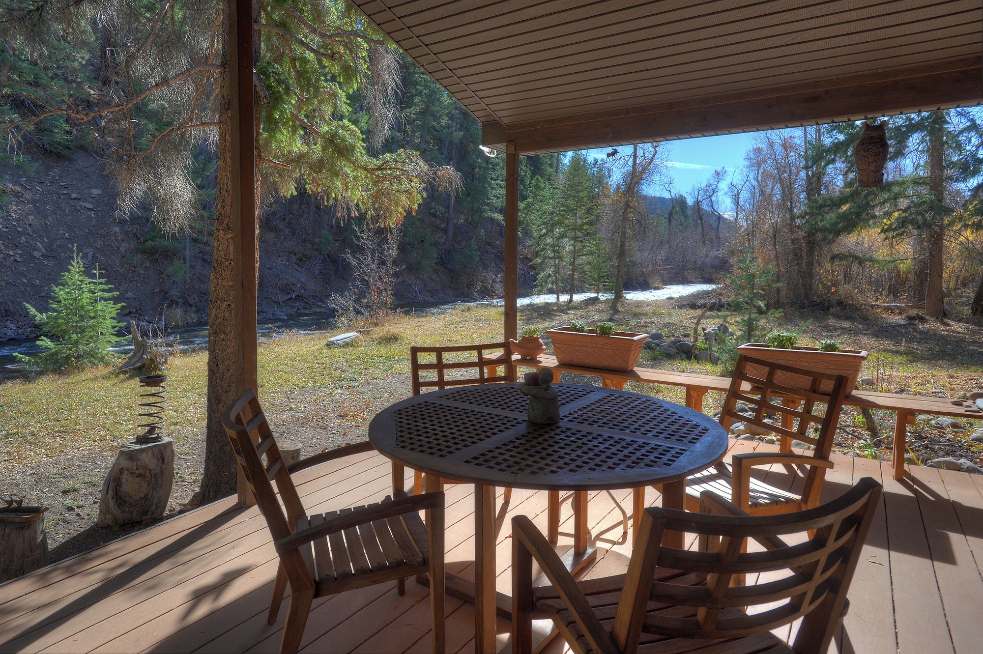 A wooden deck with a round table and chairs, overlooking a forested area with a river and a clear sky.