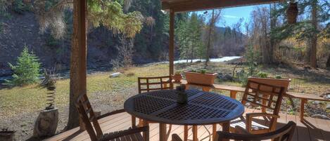 A wooden deck with a round table and chairs, overlooking a forested area with a river and a clear sky.