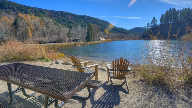 Un espace de pique-nique au bord du lac, avec une table en métal et deux chaises Adirondack, entouré d’un feuillage automnal et sous un ciel dégagé.