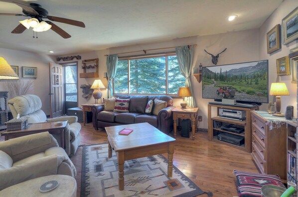 A cozy living room with a leather sofa, a wooden coffee table, and a television. There are two lamps, a rug, and a bookshelf with books and decorative items.