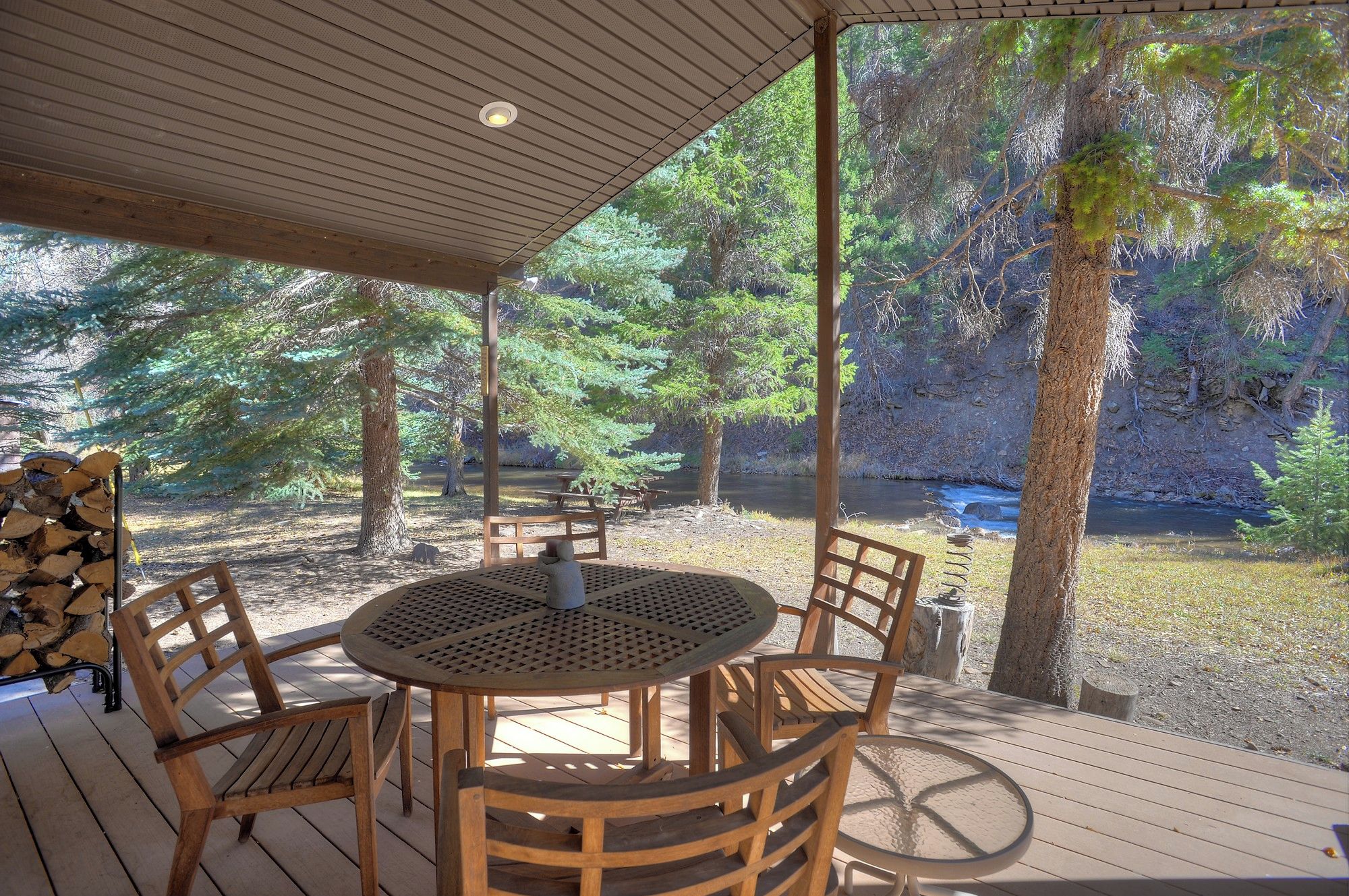 A covered outdoor seating area with a round table and chairs, surrounded by trees and a view of a river.