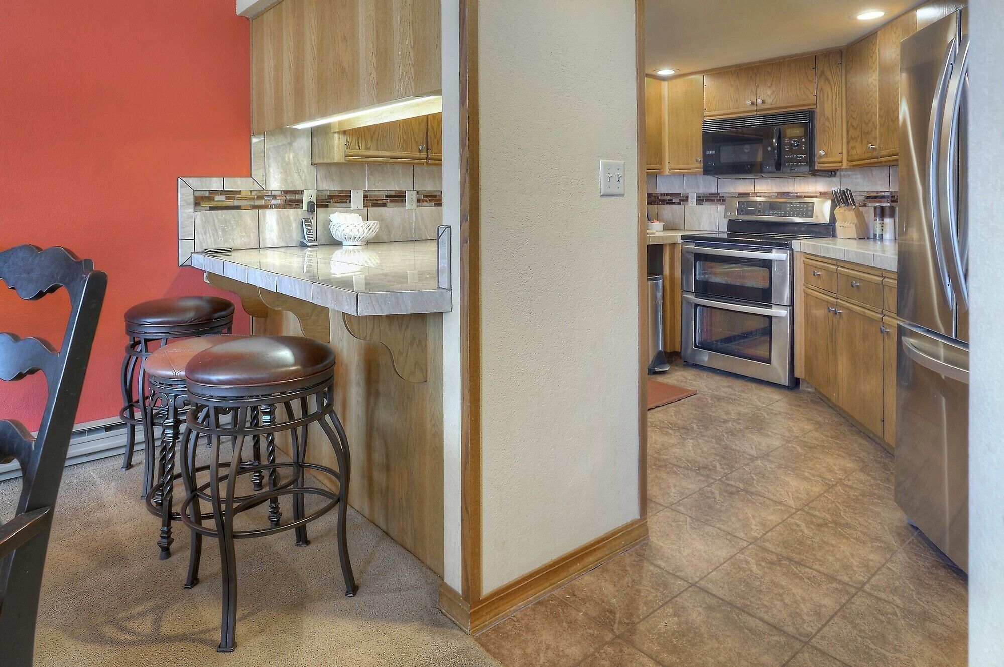 A compact kitchen with wooden cabinets, a stainless steel refrigerator, a microwave, and a double oven. There are two stools at a bar counter.