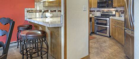 A compact kitchen with wooden cabinets, a stainless steel refrigerator, a microwave, and a double oven. There are two stools at a bar counter.