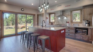 Een moderne keuken met een centraal eiland, barstoelen, een roestvrijstalen fornuis en een groot raam met uitzicht op een bebost gebied.