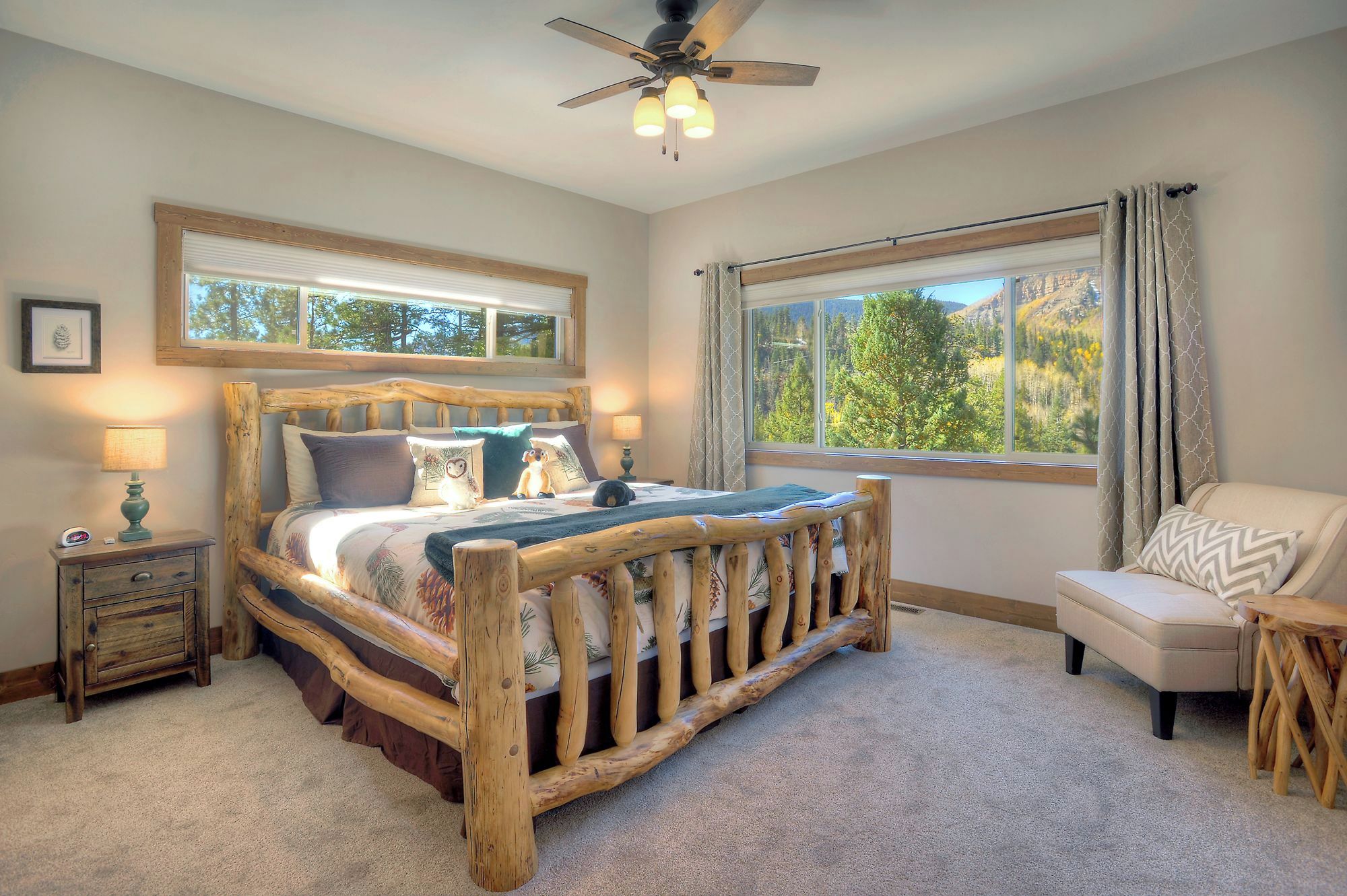 A cozy bedroom with a wooden bed frame, a nightstand with a lamp, a chair, and a large window with a view of trees.