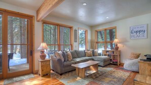 A spacious living room with a large gray sectional sofa, wooden coffee table, and a rug. There are multiple windows providing views of the surrounding forest. - Creekside Retreat - Waterfront Home 20 Minutes from Downtown Durango (Durango)
