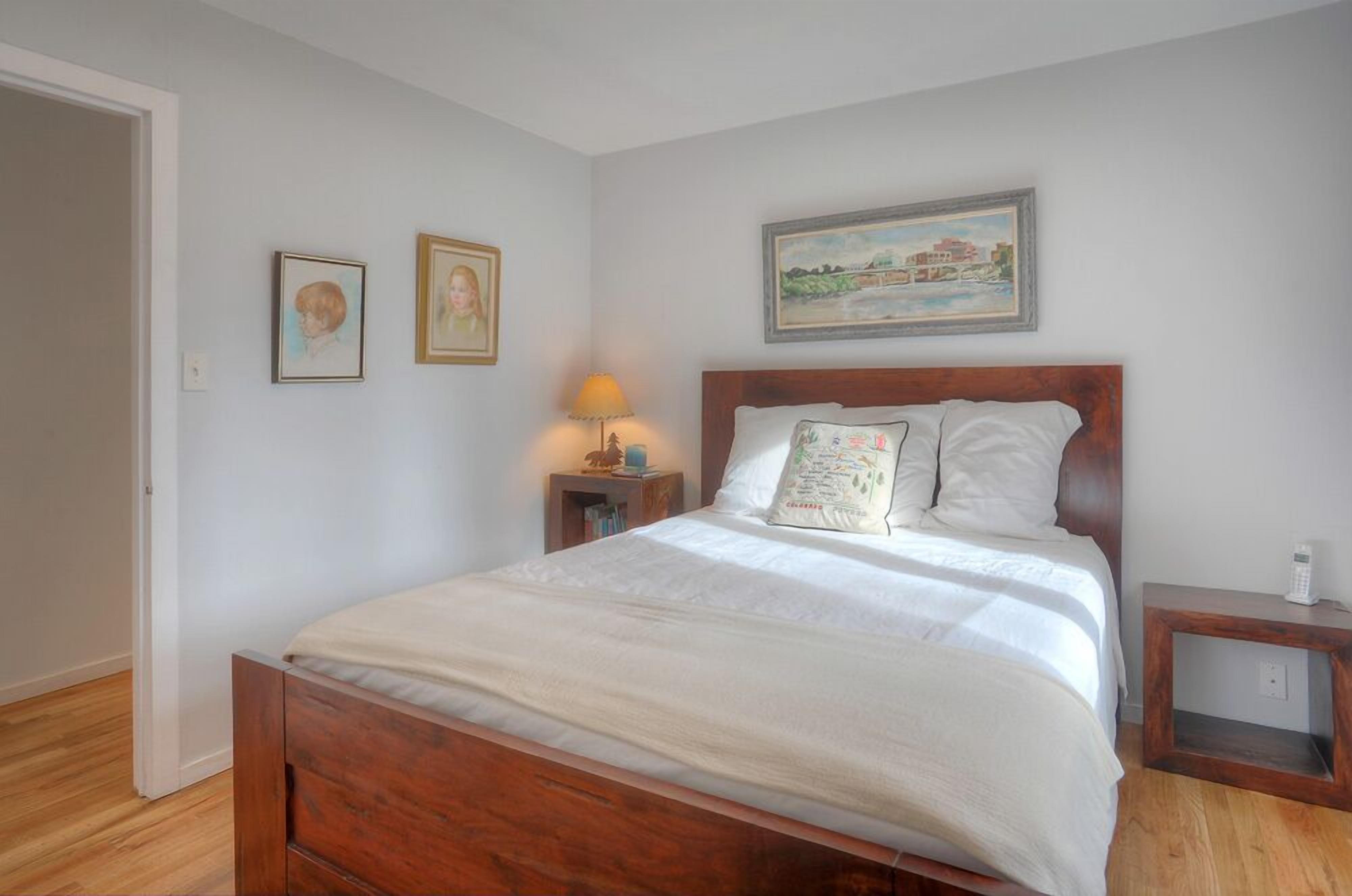 Un dormitorio con cama de madera, ropa de cama blanca, una mesita de noche con lámpara, y dos cuadros enmarcados en la pared.