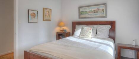Un dormitorio con cama de madera, ropa de cama blanca, una mesita de noche con lámpara, y dos cuadros enmarcados en la pared.