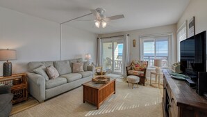 A living room with a grey sofa, wooden coffee table, and a ceiling fan. There is a TV on a wooden stand and a window with blinds. - Afterdeck 104 - Two Bedroom, Two Bath Oceanfront Condo (Garden City Beach)