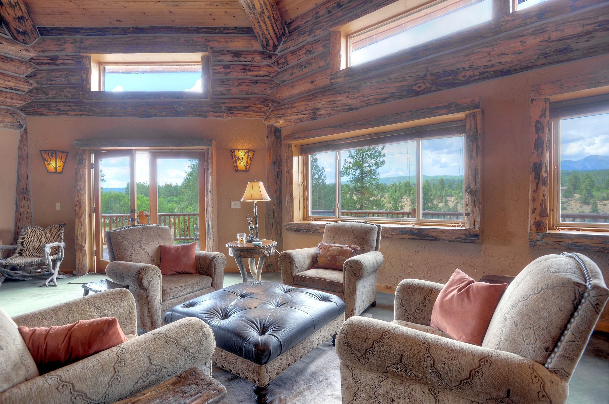 A log cabin interior with a fireplace, multiple seating areas, and large windows offering scenic views.