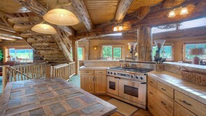 L’intérieur d’un chalet rustique est agrémenté d’un escalier en bois, d’une grande table à manger, d’une cuisine avec un poêle et de grandes fenêtres offrant une vue sur l’extérieur.