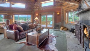 L’intérieur d’un chalet rustique, avec un canapé en cuir, une table basse en bois, une cheminée et de grandes fenêtres donnant sur le paysage extérieur.