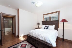 A bedroom with a wooden bed, a nightstand, a lamp, a window with blinds, and a door leading to a bathroom.