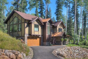 A two-story house with a stone foundation, large windows, and a brown roof, surrounded by a forested area.