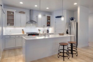 A modern kitchen with white cabinetry, a stainless steel range hood, a built-in oven, a sink, and a refrigerator. There are two stools at the island.