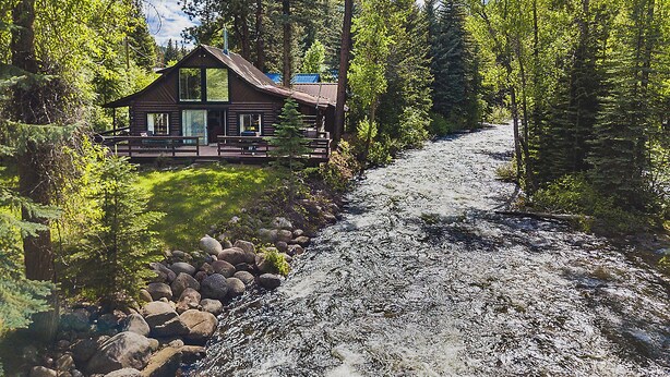 A rustic cabin with a large front porch, surrounded by dense forest and a flowing river.