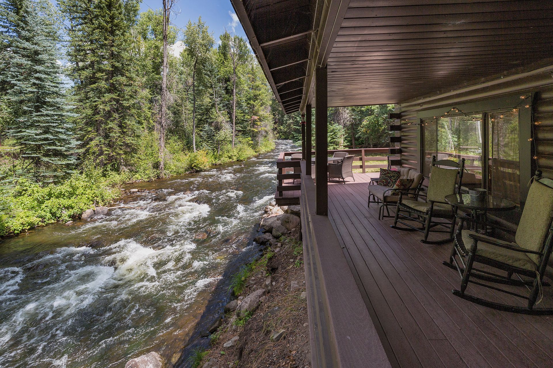 A wooden deck with outdoor seating overlooking a flowing river, surrounded by dense forest.