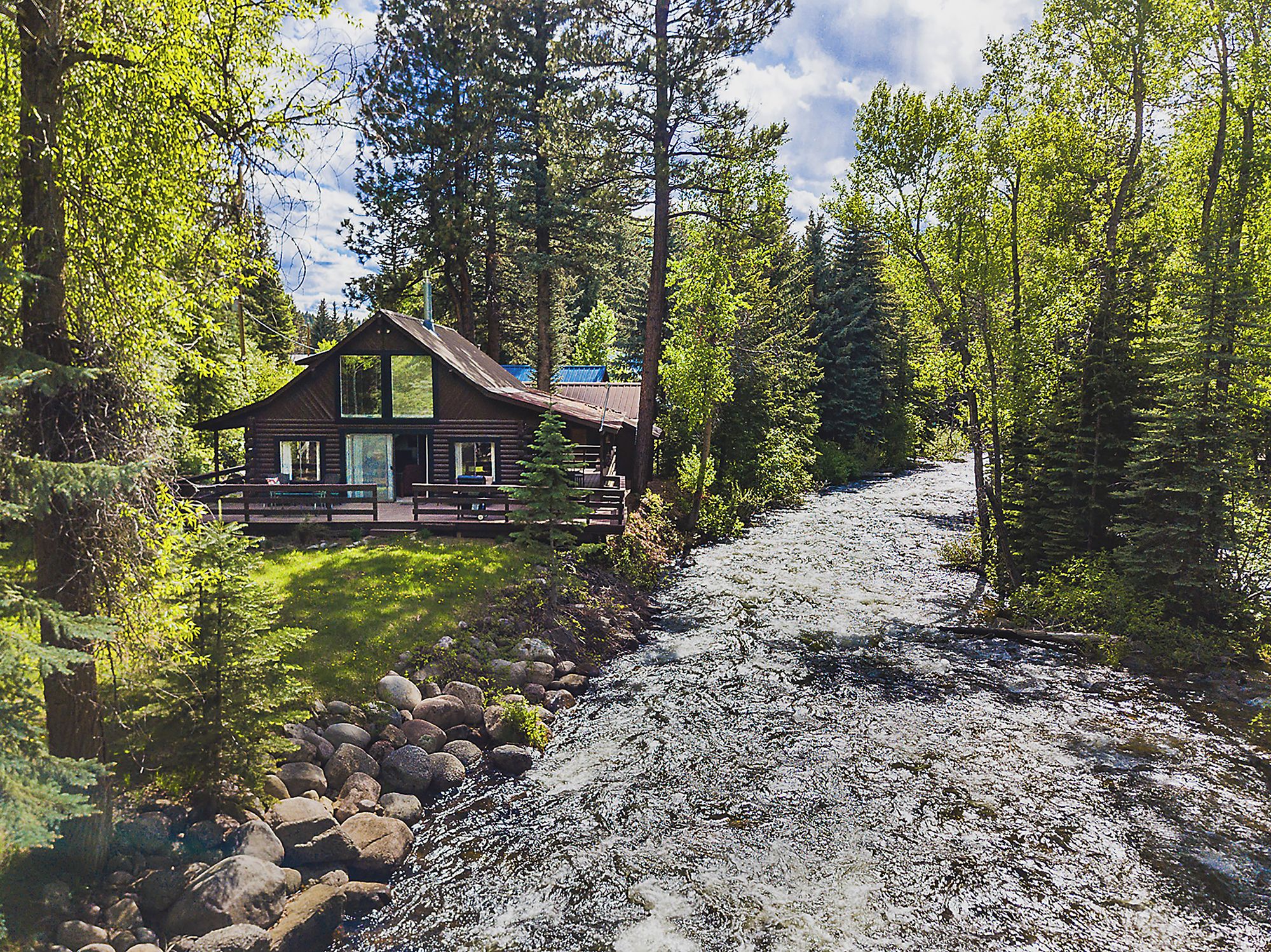 A rustic cabin with a large front porch, surrounded by dense forest and a flowing river.