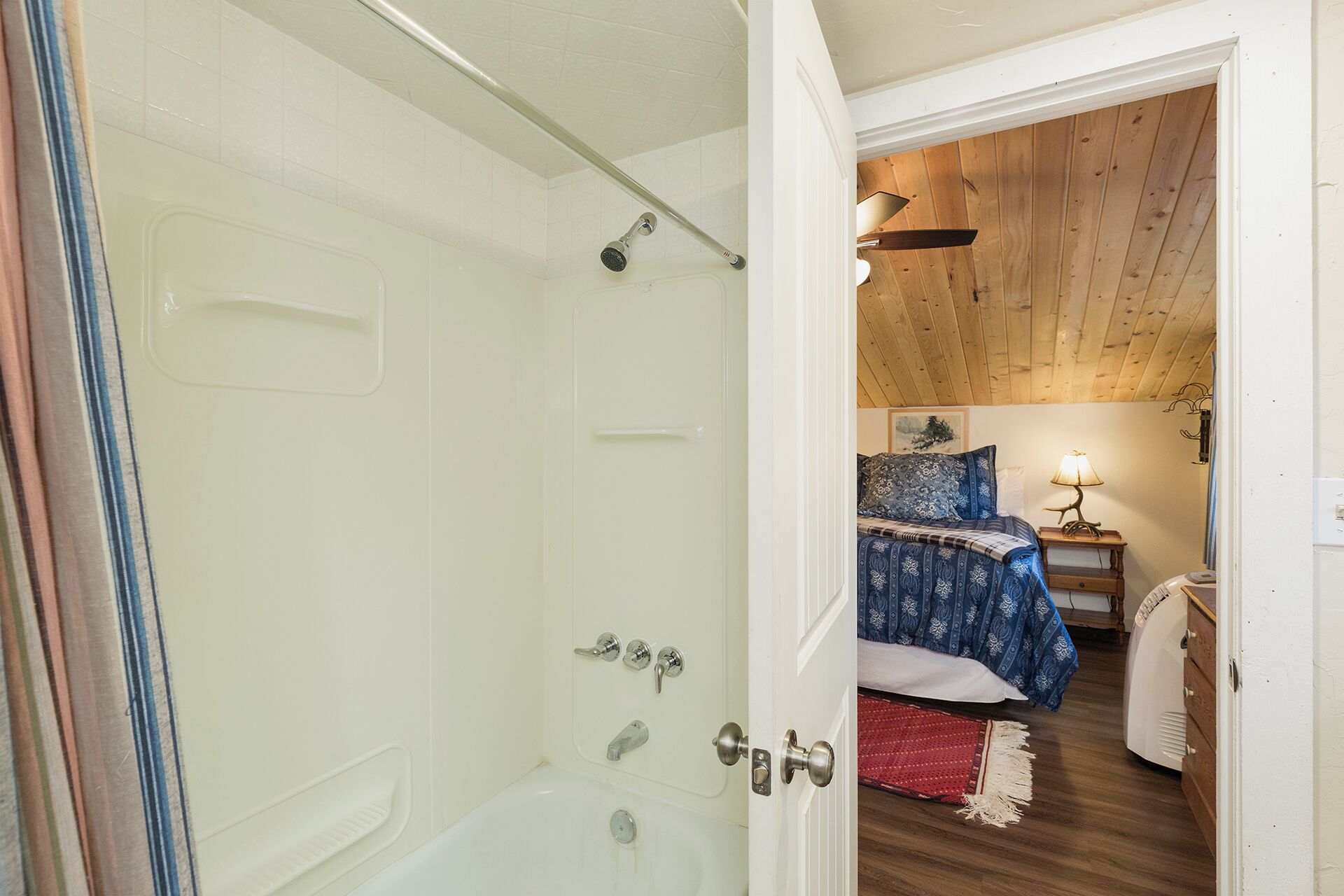 A bathroom with a bathtub and shower, a wooden ceiling, and a bedroom with a bed and nightstand.