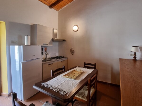 A compact kitchen with a refrigerator, stove, and sink. There is a dining table with chairs and a lamp on a wooden side table.