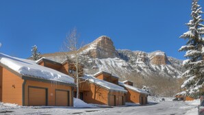 A row of wooden cabins with snow-covered roofs, set against a backdrop of snow-capped mountains under a clear blue sky.