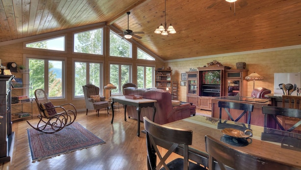 A spacious living room with large windows, wooden floors, and a high ceiling with a fan.