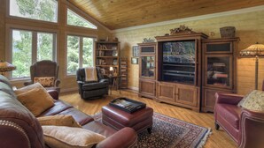 A spacious living room with leather sofas, a wooden ceiling, large windows, a bookshelf, and a television.