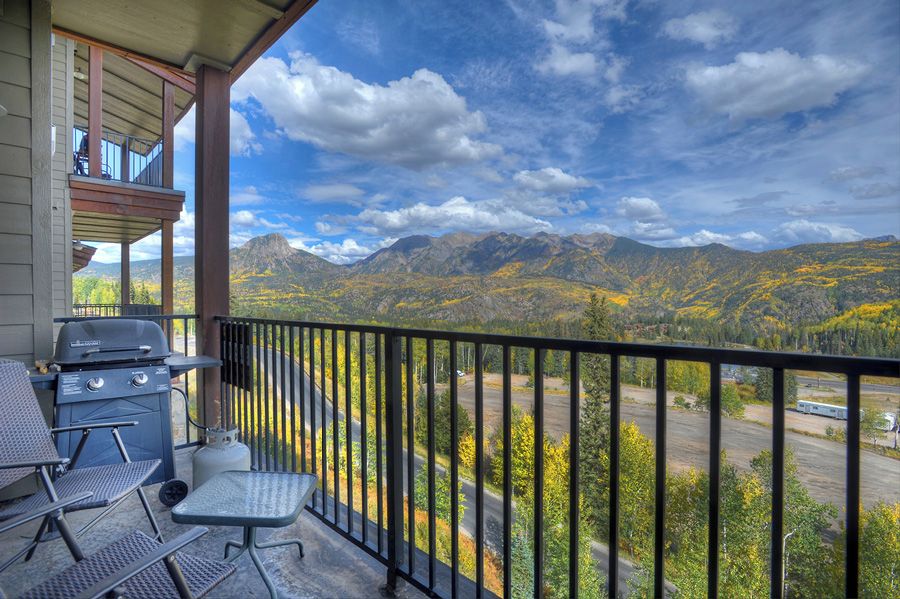 A balcony with a grill, chairs, and a table overlooking a mountainous landscape.