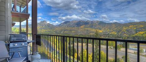 A balcony with a grill, chairs, and a table overlooking a mountainous landscape.