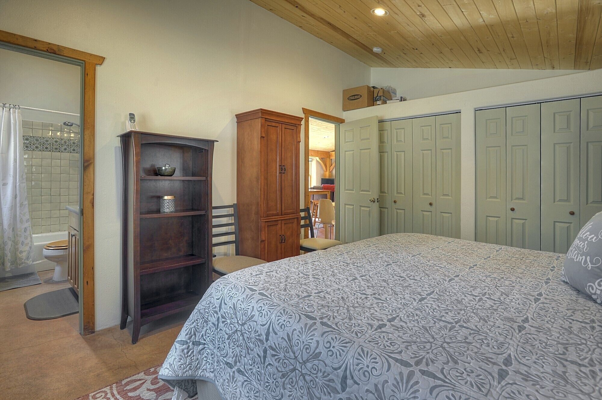 A bedroom with a bed, a wooden cabinet, a bathroom with a shower, and a set of green doors.