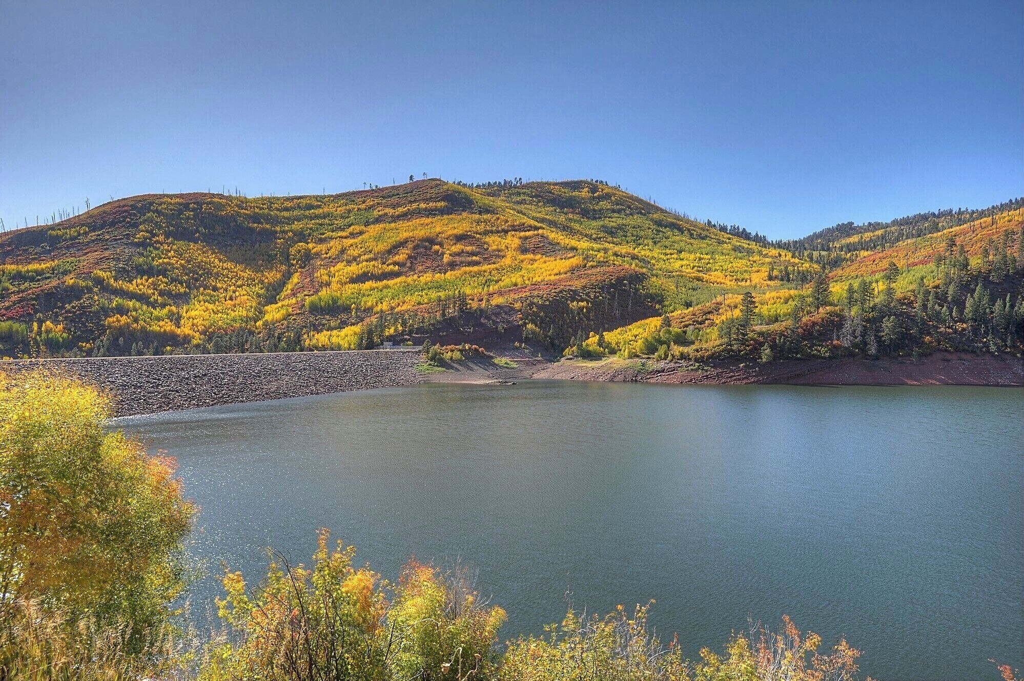 A lake surrounded by hills with autumn foliage, a stone dam, and clear skies.