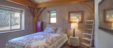 A bedroom with a wooden ceiling, a bed with a floral quilt, a nightstand with a lamp, a wooden ladder shelf, and a window with a view of trees.