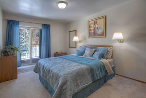 Quarto com uma cama grande, mesa de cabeceira, luminária, pintura e vista do exterior por uma porta de correr.