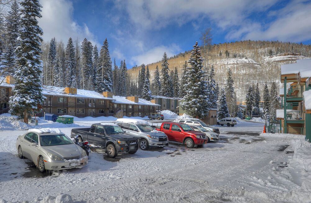 A snow-covered parking lot with several cars and a building with multiple windows and chimneys.
