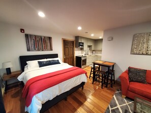 Une chambre à coucher avec un lit, une couverture rouge et blanche, un plancher en bois, une petite table avec deux tabourets, un canapé rouge et un coin cuisine équipé d’un four à micro-ondes et d’une cuisinière.
