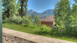 A wooden cabin with a sloped roof, surrounded by lush greenery and mountains in the background.