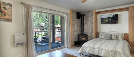 A bedroom with a bed, a ceiling fan, a fireplace, and a balcony with a view of the outdoors.