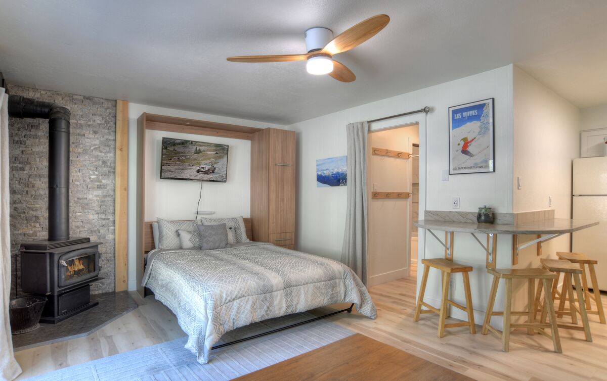 Un dormitorio con cama, ventilador de techo de madera, chimenea y zona de comedor con taburetes.