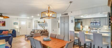 Cocina moderna con una isla central, armarios blancos y una zona de comedor con mesa y sillas de madera.