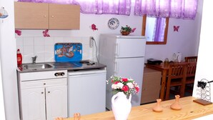 A compact kitchen with white cabinets, a sink, a refrigerator, and a dining area with a wooden table and chairs. - Marianthi Studios Stegna (Rhodes)