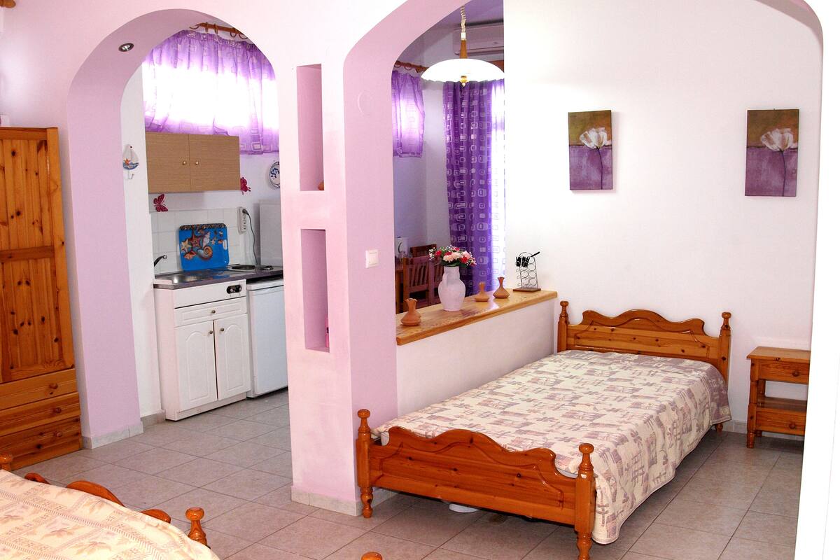 Un dormitorio con una cama de madera, una mesita de noche pequeña y una mesita auxiliar. Hay una ventana con cortinas moradas y un cuadro en la pared.